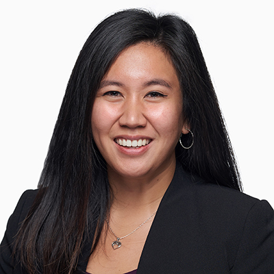 A woman with long dark hair, wearing a black blazer, hoop earrings, and a necklace, smiles at the camera against a plain white background.