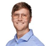 Smiling man with light brown hair, wearing glasses and a light blue collared shirt, posed against a plain white background.