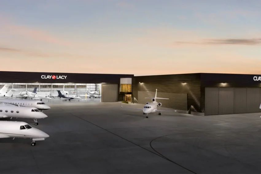 Several private jets are parked outside a modern hangar with the sign "Clay Lacy" at sunset.