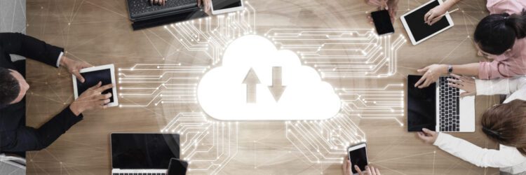 A cloud computing graphic showing various cloud types is displayed on a table surrounded by people using laptops, tablets, and smartphones.
