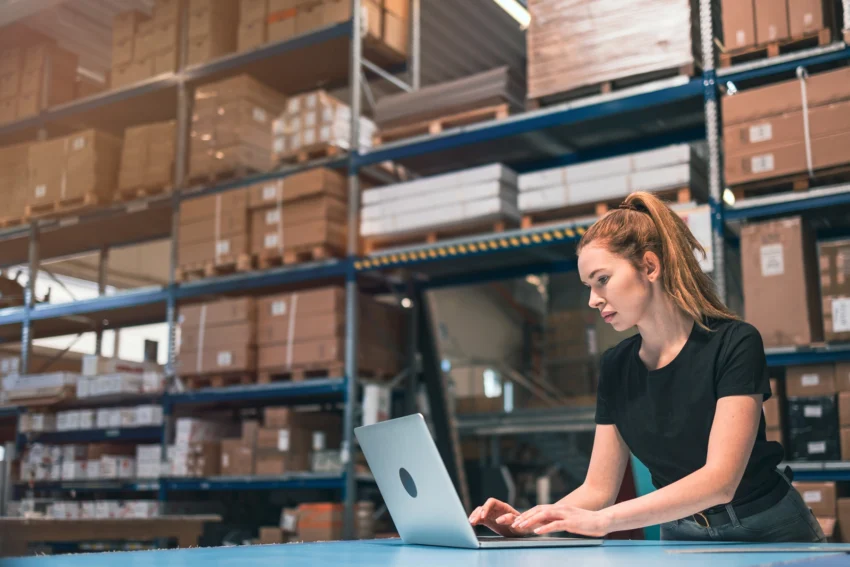 A woman using a laptop in a warehouse, surrounded by shelves filled with boxes and packages.