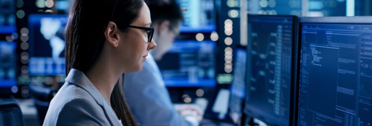 A woman wearing glasses works at a computer with multiple monitors displaying code, possibly advancing her OnBase Admin Career; another person is visible working in the background in a tech office setting.