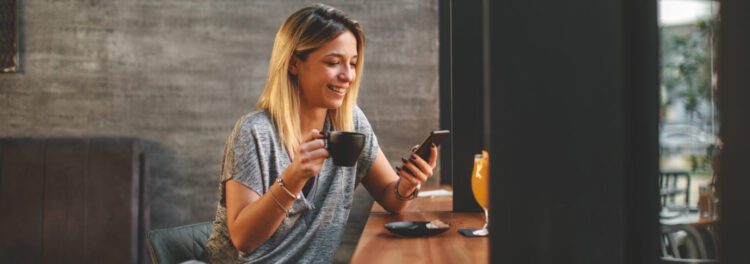 A woman sits at a café table, smiling while looking at her phone and holding a coffee cup, bridging the digital gap as she enjoys a glass of juice and a small plate in front of her.