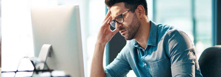 A man wearing glasses and a denim shirt sits at a desk in a modern office, looking stressed as he works on his computer, possibly searching for workflow tips to improve his productivity.