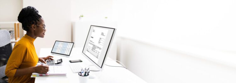 A woman sits at a desk using a computer, calculator, and documents, working with spreadsheets and financial data to manage cloud savings efficiently.