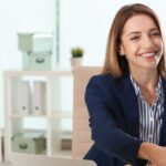 Three people in business attire sit at a table in an office; a woman in a navy blazer is smiling and shaking hands with a man across from her, reflecting the positive impact of HR transformation.