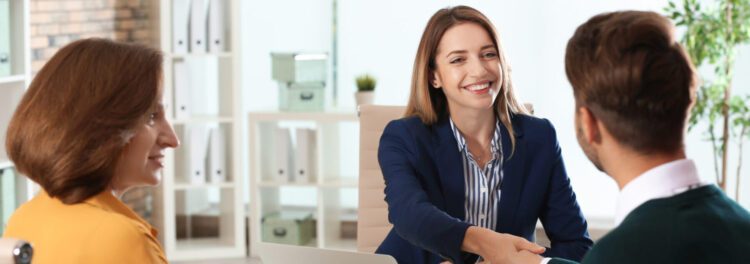 Three people in business attire sit at a table in an office; a woman in a navy blazer is smiling and shaking hands with a man across from her, reflecting the positive impact of HR transformation.