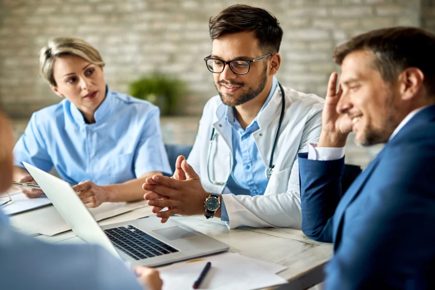 Three healthcare professionals and one person in a suit have a discussion around a table, with a laptop open in front of them.