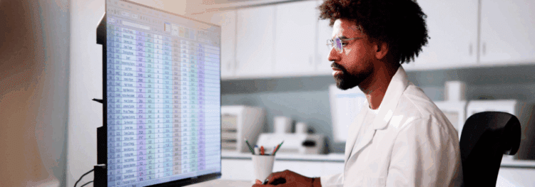 A scientist in a lab coat analyzes healthcare auditing data on a large computer screen displaying a spreadsheet with rows and columns in a laboratory setting.