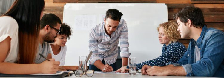 Five people sit around a table in a meeting room, discussing AP goals and documents, with one person standing and writing on a paper; a whiteboard is in the background.