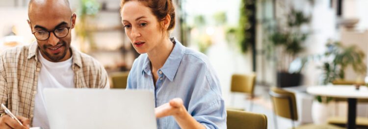 Two people sit at a table looking at a laptop screen, discussing data literacy while taking notes, in a modern, brightly lit indoor setting.