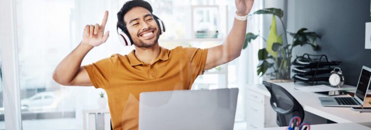 A man wearing headphones smiles and raises his arms while sitting at a desk with a laptop, celebrating a successful onbase rpa project in a bright, modern office.