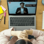 A person sits at a desk watching a webinar on a laptop, surrounded by stationery, a notebook, sticky notes, a potted plant, and a cup of coffee, focused on upskilling and reskilling for career growth.