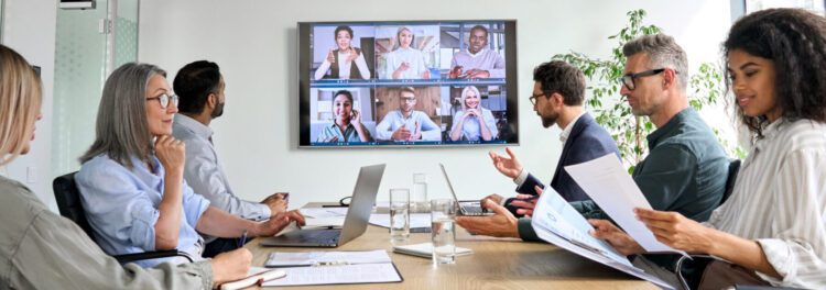 A group of people sit around a conference table with laptops and papers, participating in a video call focused on sustainable digital transformation with colleagues shown on a large screen.