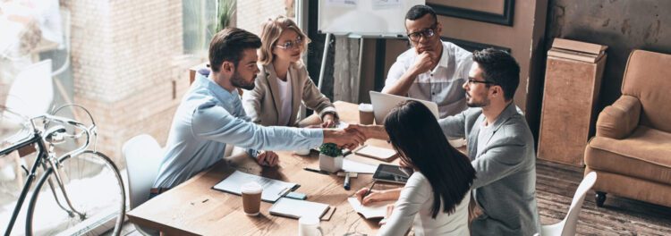 Five people sit around a table in an office, two of them shaking hands, suggesting a business meeting or agreement focused on breaking down silos. Laptops, papers, and coffee cups are scattered across the table.