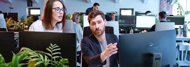 Two colleagues in a modern office discuss work for a data migration company while looking at a computer screen. Other employees are visible in the background at their desks.