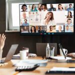 A group of people in a conference room wave at colleagues on a large monitor during a video call, discussing open source change management, with laptops and documents on the table.