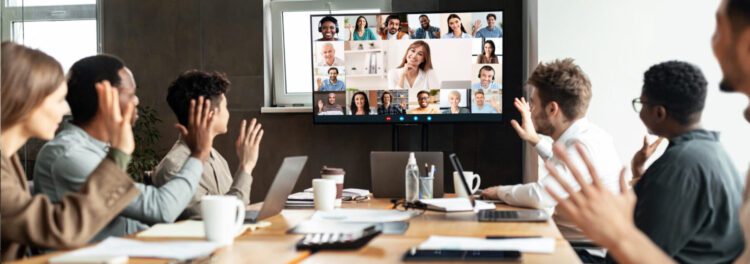 A group of people in a conference room wave at colleagues on a large monitor during a video call, discussing open source change management, with laptops and documents on the table.