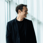 Two men in business attire walk down a bright hallway, engaged in conversation about IT trends 2025. One gestures with his hands while the other smiles and listens intently.