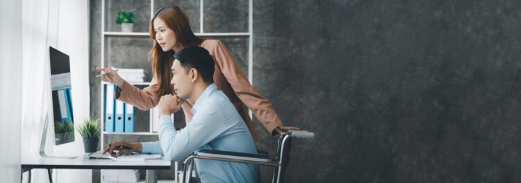 A woman stands beside a man in a wheelchair at a desk, both looking at a computer screen in a modern office setting as they discuss intelligent automation use cases.
