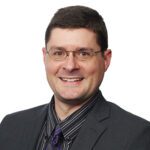 Man wearing glasses, a dark suit, a black and gray striped shirt, and a purple striped tie, smiling at the camera against a plain white background.