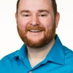 A man with short brown hair and a beard smiles at the camera. He is wearing a bright blue collared shirt against a plain white background.
