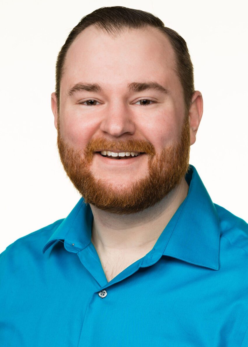 A man with short brown hair and a beard smiles at the camera. He is wearing a bright blue collared shirt against a plain white background.