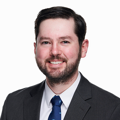 A man with short dark hair and a beard, wearing a dark suit, white shirt, and blue tie, poses against a plain white background.