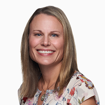 Woman with straight blond hair smiling, wearing a floral blouse and a necklace, photographed against a plain white background.