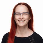 A woman with long red hair, wearing glasses and a black sweater, smiles at the camera against a plain white background.