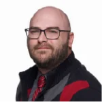 A man with glasses, a beard, and a bald head wears a striped sweater and a red patterned tie, posing against a plain white background.
