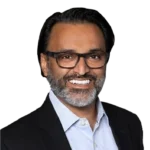 A man with dark hair and a beard, wearing glasses, a dark suit jacket, and a light blue shirt, smiling at the camera against a plain background.