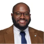 A man with glasses, a beard, and a bald head, wearing a brown jacket, white shirt, and dark tie, smiles at the camera against a white background.
