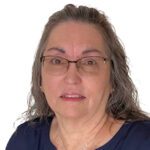 Woman with long gray hair and glasses wearing a navy blue top, facing the camera against a white background.