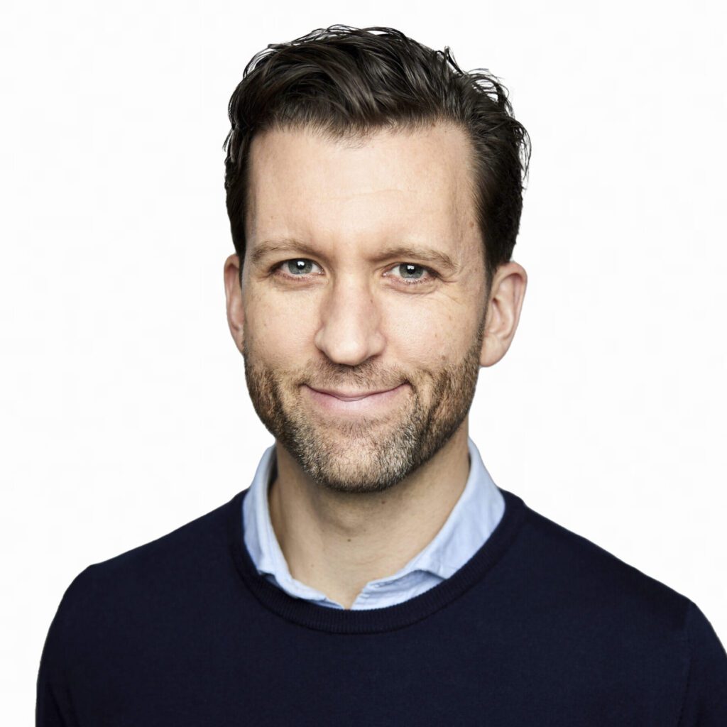 Man with short brown hair and beard, wearing a navy sweater over a light blue collared shirt, smiling at the camera against a plain white background.