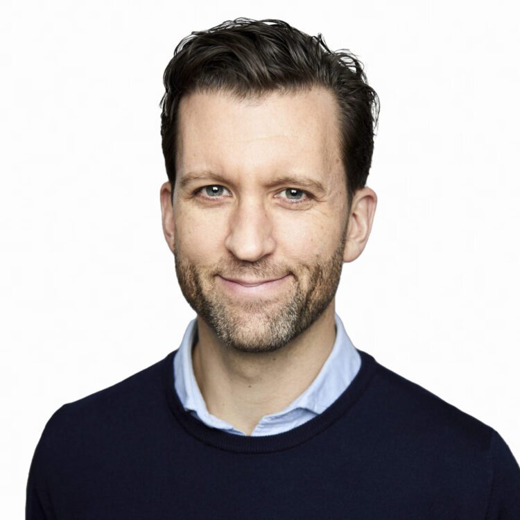 Man with short brown hair and beard, wearing a navy sweater over a light blue collared shirt, smiling at the camera against a plain white background.