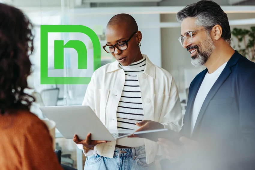 Three people in an office setting have a discussion; one holds a laptop, another a tablet. A green logo resembling an "n" inside a square is displayed on glass behind them.