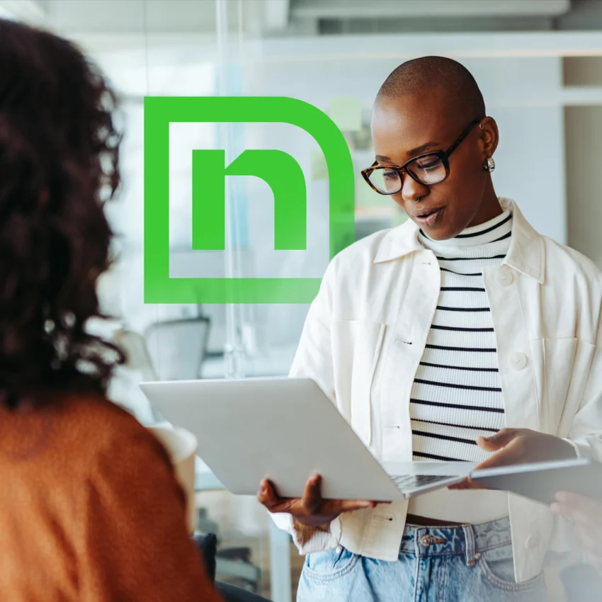 Two people stand indoors, one holding a laptop and wearing glasses, with a stylized green “n” logo on a glass wall in the background.