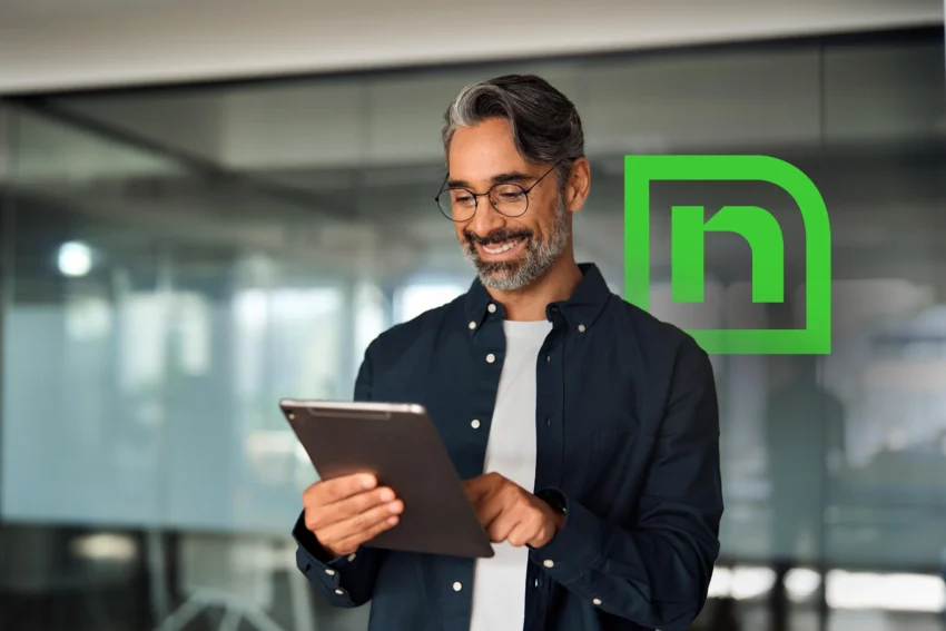 A man with glasses and gray hair stands indoors, smiling while using a tablet. A green stylized logo is displayed on the glass wall behind him.