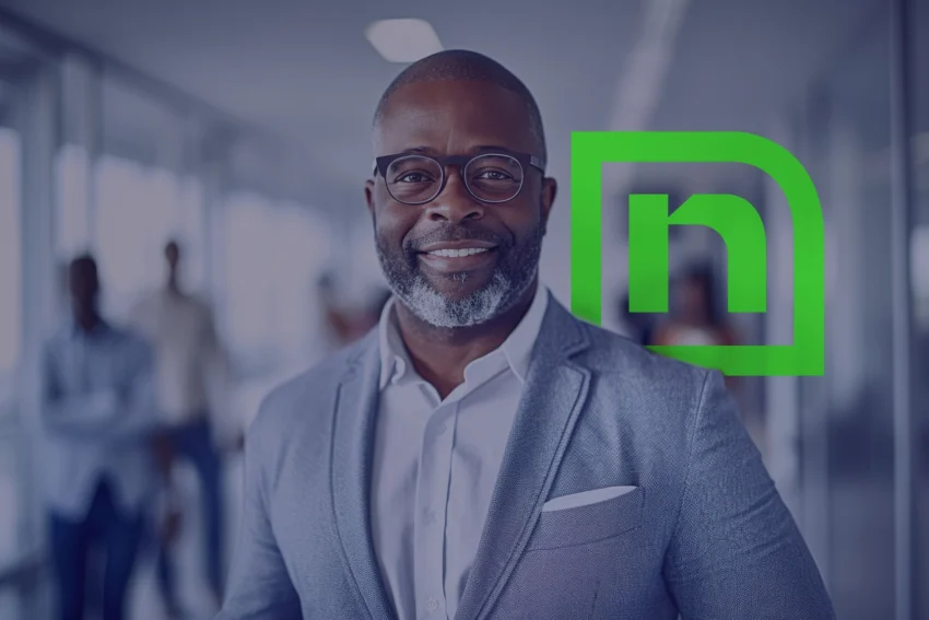 A man in a gray suit and glasses stands smiling in an office hallway. A green stylized "n" logo is displayed beside him.