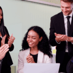 Five professionally dressed people are sitting and standing around a table, smiling and clapping, with a laptop and tablets visible—celebrating successful process orchestration use cases in a modern office setting.