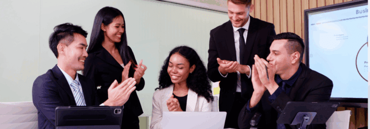 Five professionally dressed people are sitting and standing around a table, smiling and clapping, with a laptop and tablets visible—celebrating successful process orchestration use cases in a modern office setting.