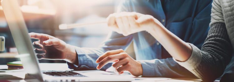 Two people sitting at a desk work together on a laptop, discussing RPA vs. API; one person points at the screen with a pencil while the other gestures toward the keyboard.