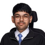 A young man with short dark hair, glasses, and a trimmed beard, wearing a white shirt, blue tie, and black jacket, sits in a wheelchair against a plain white background.