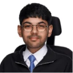 A young man with short black hair and glasses, wearing a black jacket, white shirt, and blue tie, sits in a wheelchair against a white background.