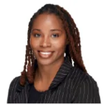 A woman with long, braided hair, wearing a black pinstripe blazer, smiles at the camera against a white background.