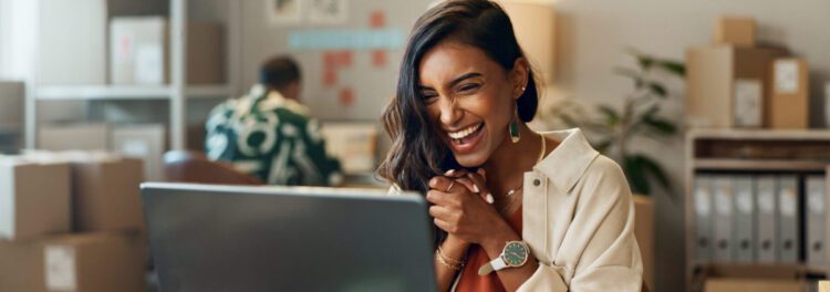 A woman sits at a desk smiling and clasping her hands while looking at a laptop, exploring low code technology, with office supplies and boxes in the background.