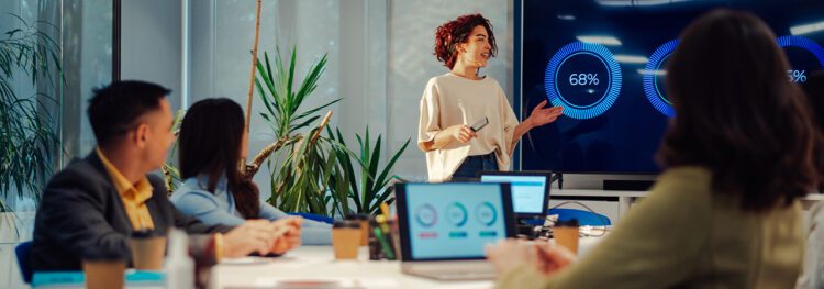 A woman presents data visuals on a large screen to three seated colleagues in a modern office meeting room, facilitating effective change management communication.