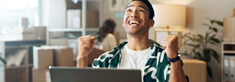A person sits at a desk in front of a laptop, smiling and raising clenched fists in excitement—perhaps celebrating the low code benefits that make work easier. Boxes and office supplies are visible in the background.