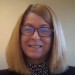 A woman with straight, shoulder-length blonde hair and glasses smiles at the camera. She is wearing a dark patterned top and is indoors with a neutral background.